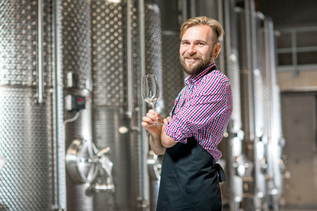Portrait of a handsome wine maker in working apron with the wine glass at the manufacture with metal tanks for wine fermentation. Wine production at the modern factoryの写真素材