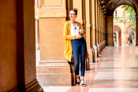 Young female student dressed casually standing with books in the famous arched galleries in Bologna city in Italy. Bologna is student city and home to the oldest university in the worldの写真素材