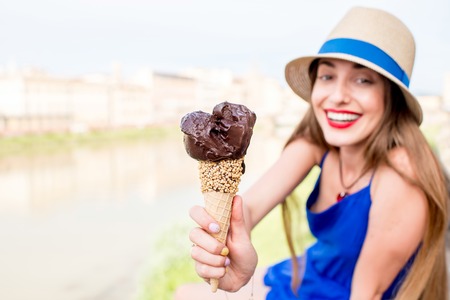 Young female tourist in blue dress holding chocolate ice cream in waffle cone sitting near the river in Florence city. Image with small depth of field focused on ice creamの写真素材