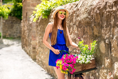 Beautiful woman in blue dress and hat standing with retro bicycle full of flowers in the old italian townの写真素材
