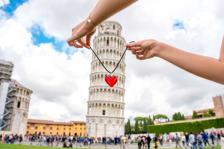 Holding red pendant in form of the heart above the famous leaning tower in Pisa old town in Italy.の写真素材