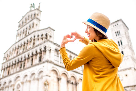 Young female traveler in yellow sweater and hat showing heart shape in front of San Michele basilica...