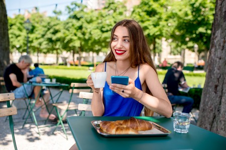 Beautiful woman in the blue dress sitting at the cafe with coffee and croissant using mobile phone outdoors at the park.の写真素材