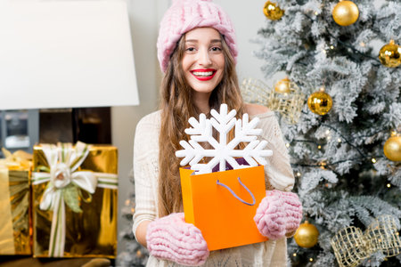 Young smiling woman in knitted hat and gloves holding shopping bag with snow flake standing in decorated home interior with Christmas tree. Winter holiday shopping conceptの写真素材