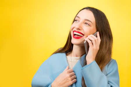 Young happy woman in the blue coat talking with mobile phone on the yellow wall backgroundの写真素材