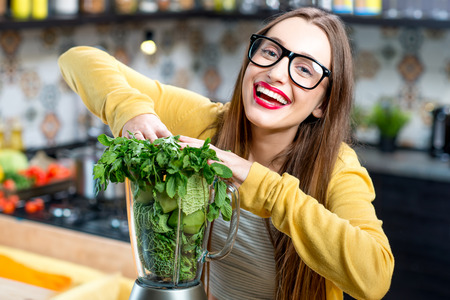 Young smiling woman pushing fresh greens into the blender in the kitchen at home. Healthy vegetarian smoothie for weight loss and detoxの写真素材