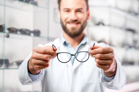 Handsome ophthalmologist holding eyeglasses for a try out. Optometrist offering to wear a pair of glasses. Image with small deph of field focused on the hands and glassesの写真素材