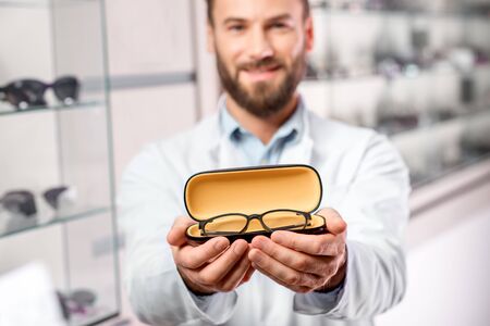 Handsome ophthalmologist holding eyeglasses for a try out. Optometrist offering to wear a pair of glasses. Image with small deph of field focused on the hands and glassesの写真素材