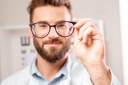 Handsome ophthalmologist looking through the glasses on the eye chart background in the cabinet. Image with small deph of field focused on hands and glassesの写真素材