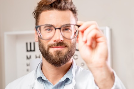Handsome ophthalmologist looking through the glasses on the eye chart background in the cabinetの写真素材