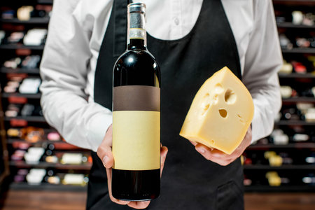Food seller holding a bottle of white wine and a pieace of gouda cheese. Choosing wine according to the type of cheese. Bottle with empty label to copy pasteの写真素材