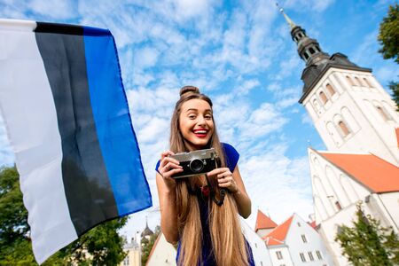 Portrait of a young female tourist with photo camera and estonian flag in front of saint Nicholas church in the old town of Tallinn. Woman having great vacations in Estoniaの写真素材