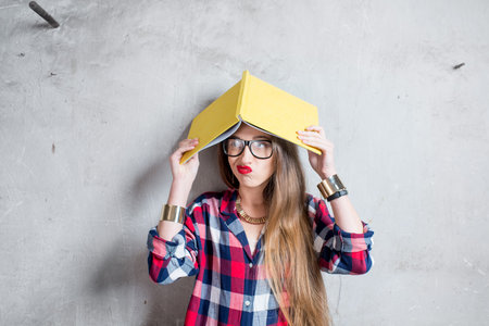 Portrait of confused student in checkered shirt with yellow book on the gray wall backgroundの写真素材