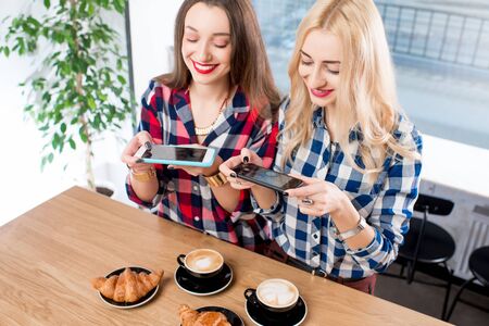 Two female friends photographing coffee with croissants on the table at the cafeの写真素材