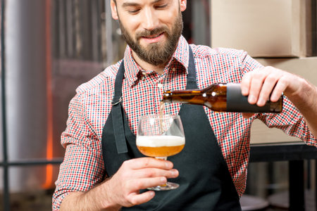 Handsome brewer in apron and shirt pouring beer into the glass at the manufacturing. Image focused...