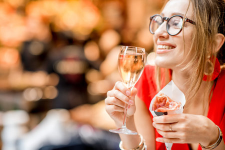 Young woman in red dress eating jamon traditional spanish dry-cured ham with glass of wine sitting...