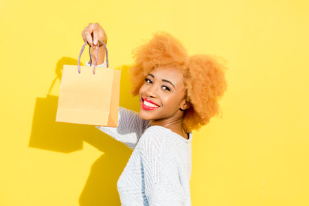 Portrait of a beautiful african woman in blue sweater standing with shopping bag on the yellow backgroundの写真素材