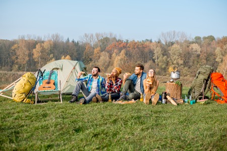 Multi ethnic group of friends having a picnic, eating pizza, sitting in a row at the camping with tent and hiking equipment near the forestの写真素材
