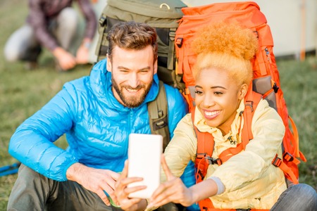 Young couple in colorful jackets with backpacks having fun with smart phone sitting on the green lawn near the camping placeの写真素材