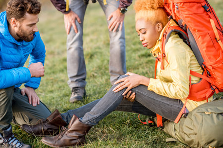 Young african woman with backpack having a trauma with knee hiking with friends outdoorsの写真素材