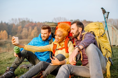 Happy friends in colorful jackets with backpacks having fun with smart phone sitting on the green lawn near the camping placeの写真素材