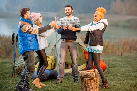 Multi ethnic group of friends dressed in sweaters celebrating with bengal fire standing together at the camping during the evening lightの写真素材