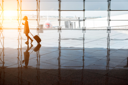 View on the aiport window with woman walking with suitcase at the departure hall during the sunset. Wide angle view with copy spaceの写真素材