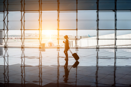 View on the aiport window with woman walking with suitcase at the departure hall during the sunset. Wide angle view with copy spaceの写真素材