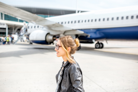 Young woman in leather jacket walking near the airplane outdoors on the airport runwayの写真素材