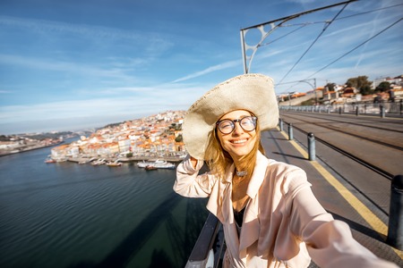 Young woman tourist in hat making selfie portrait on the cityscape background standing on the iron bridge in Porto city, Portugalの写真素材