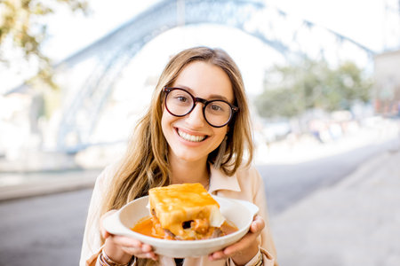 Young woman enjoying traditional portuguese meat sandwich called Francesinha, sitting at the bar with famous bridge on the background in Porto city, Portugalの写真素材