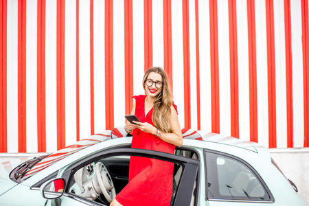 Poortrait of a young happy woman standing with phone near the car on the red wall background