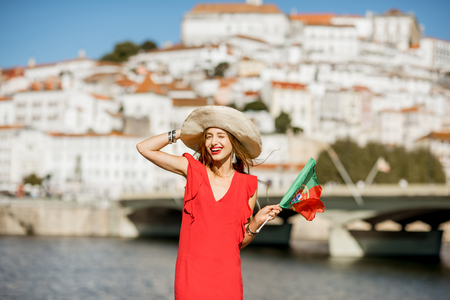 Portrait of a young woman tourist in red dress standing with portuguese flag on the Coimbra city background in central Portugalの写真素材