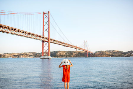 Woman in red dress enjoying landscape view on the famous iron bridge standing back on the riverside in Lisbon city, Portugal. Wide angle view with copy spaceの写真素材