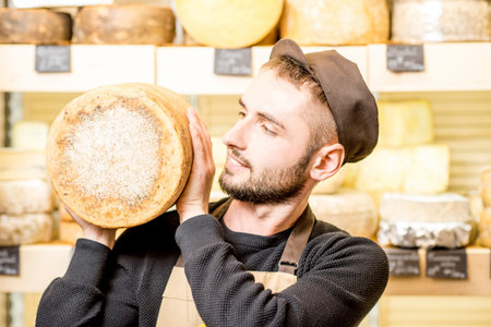 Portrait of a handsome cheese seller in uniform holding a big seasoned cheese in front of the store...