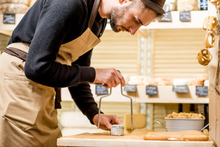 Portrait of a handsome salesman in uniform cutting cheese in front of the store showcase full of...