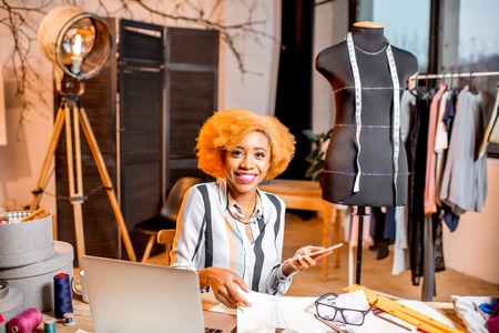 Young african fashion designer working with clothes drawings and laptop sitting at the studio interior with different tailoring toolsの写真素材