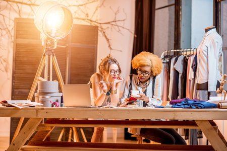 Two female multi-ethnic fashion designers working at the office with different tailoring tools and...