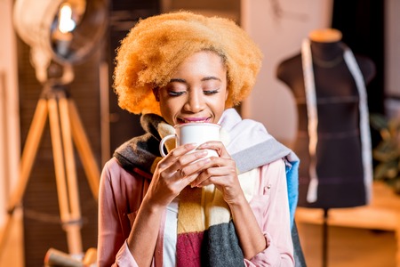 Portrait of a young african creative person with colorful scarf holding hot drink at the studio interiorの写真素材