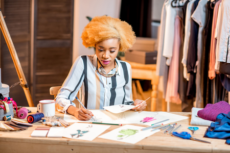 Young african fashion designer working with clothes drawings sitting at the office interior with different tailoring toolsの写真素材