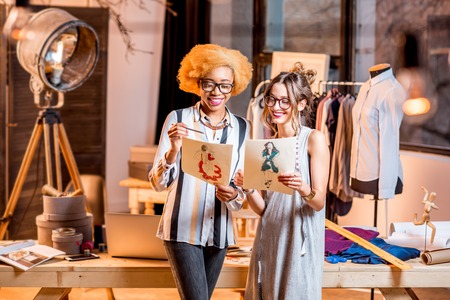 Two female multi-ethnic fashion designers working with clothes drawings standing at the beautiful office with different tailoring tools and clothesの写真素材