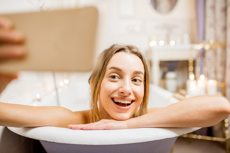 Young and beautiful woman making selfie photo with phone lying in the retro bath indoorsの写真素材