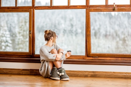Young woman in sweater sitting near the big window at the cozy wooden mountain house with beautiful landscape view during the winter timeの写真素材