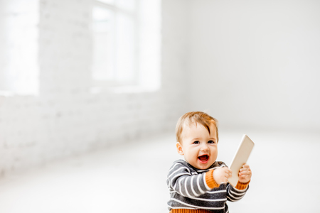 Cute and joyful baby boy playing with smartphone sitting indoors on the white floorの写真素材