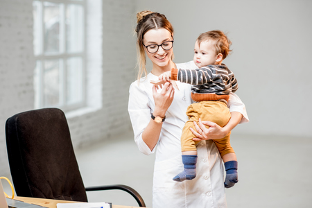 Young woman pediatrician in medical gown standing with baby boy at the white office interiorの写真素材