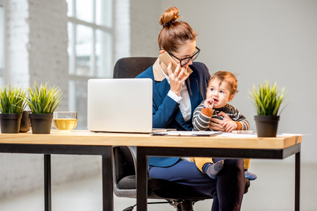 Young multitasking businessmam dressed in the suit working with laptop and documents sitting with her baby son at the officeの写真素材