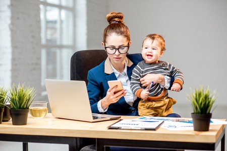 Young multitasking businessmam dressed in the suit working with laptop and documents sitting with her baby son at the officeの写真素材