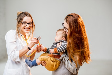 Mother with her baby boy at the young female pediatrician listening with a stethoscope standing at the white office interiorの写真素材