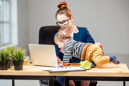 Young multitasking businessmam dressed in the suit working with laptop and documents sitting with her baby son at the officeの写真素材