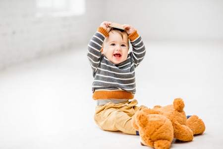 Cute and joyful baby boy playing with smartphone sitting indoors on the white floorの写真素材